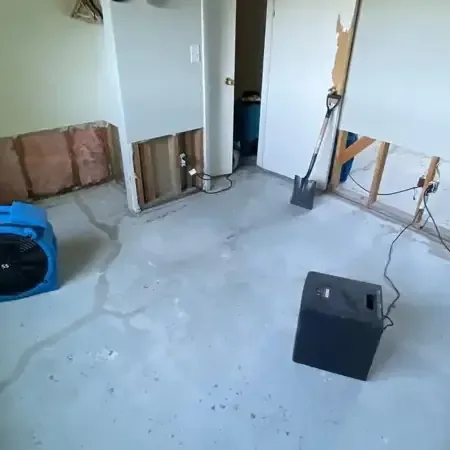 A blue air mover and a black dehumidifier on a bare concrete floor beside lower drywall cut away